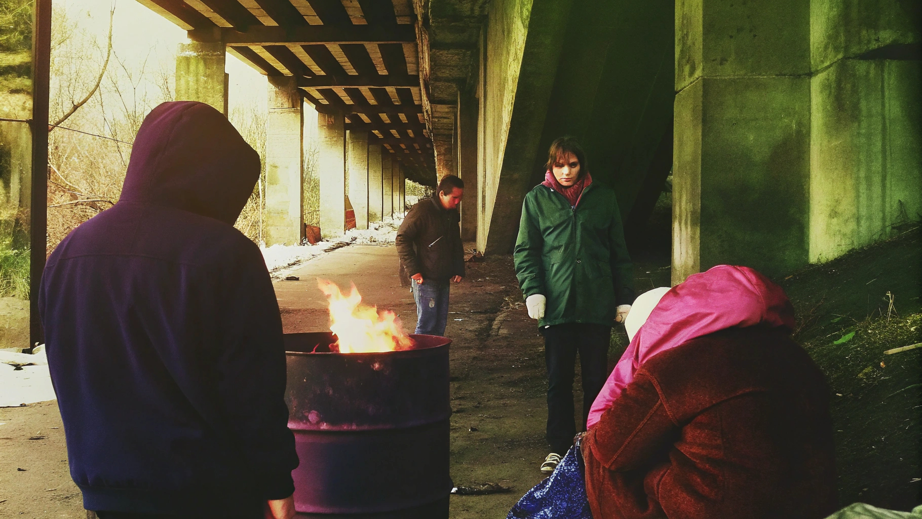 People warming by barrel fire under bridge
